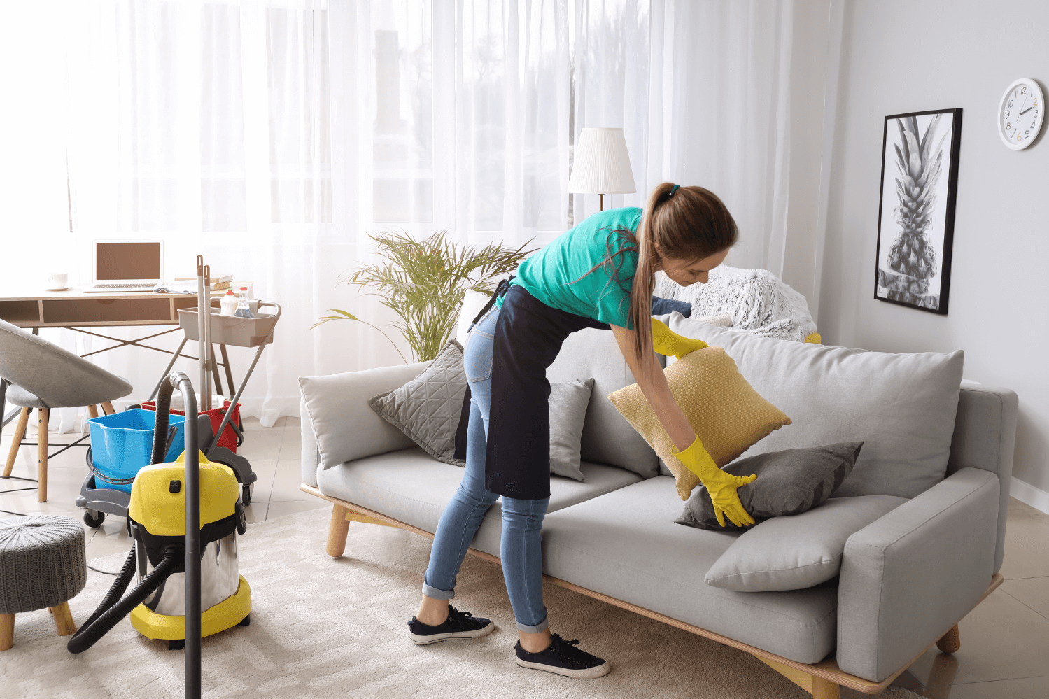 Professional cleaner arranging cushions on a sofa in a bright, modern living room with cleaning supplies in the background.