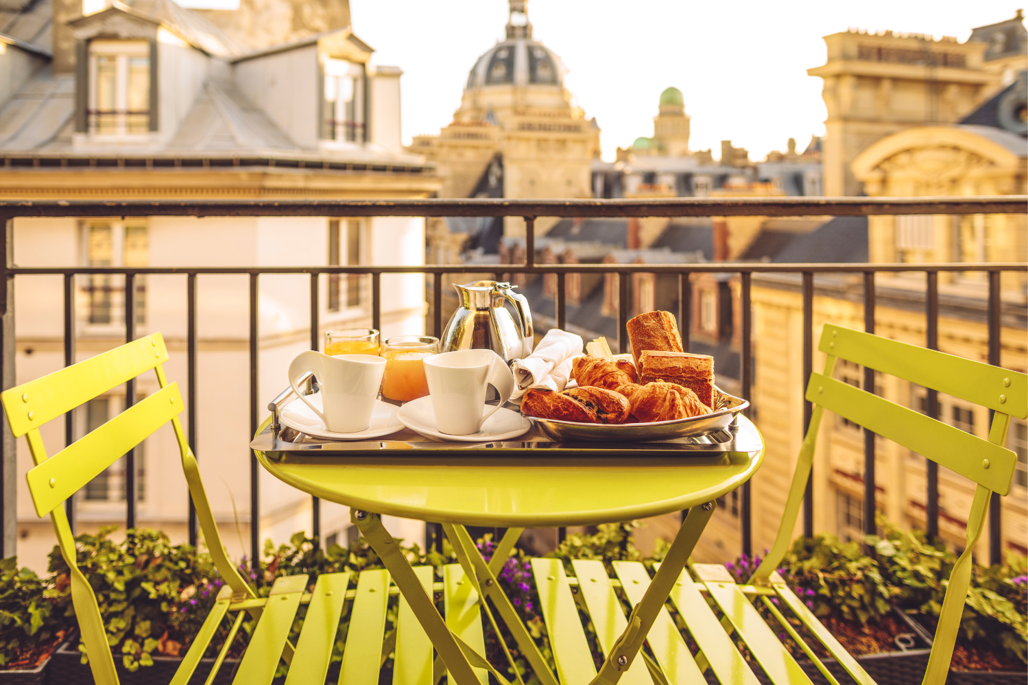 A cozy breakfast setup on a balcony with a lime-green table and chairs, featuring a tray of fresh croissants, pastries, orange juice, and coffee. The balcony overlooks a picturesque European cityscape with historic buildings and domes under the warm glow of sunlight.