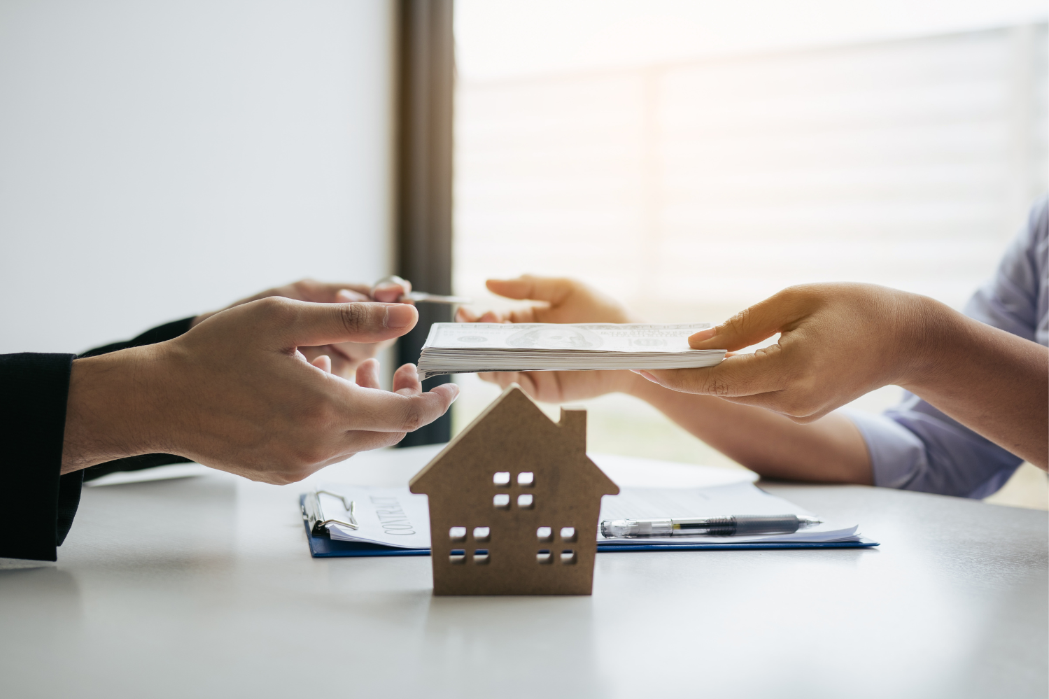 A close-up image of a real estate transaction taking place. One person is handing over a stack of money while the other exchanges keys. In the foreground, a small wooden house model sits on a table alongside a contract and a pen, symbolizing a property deal.