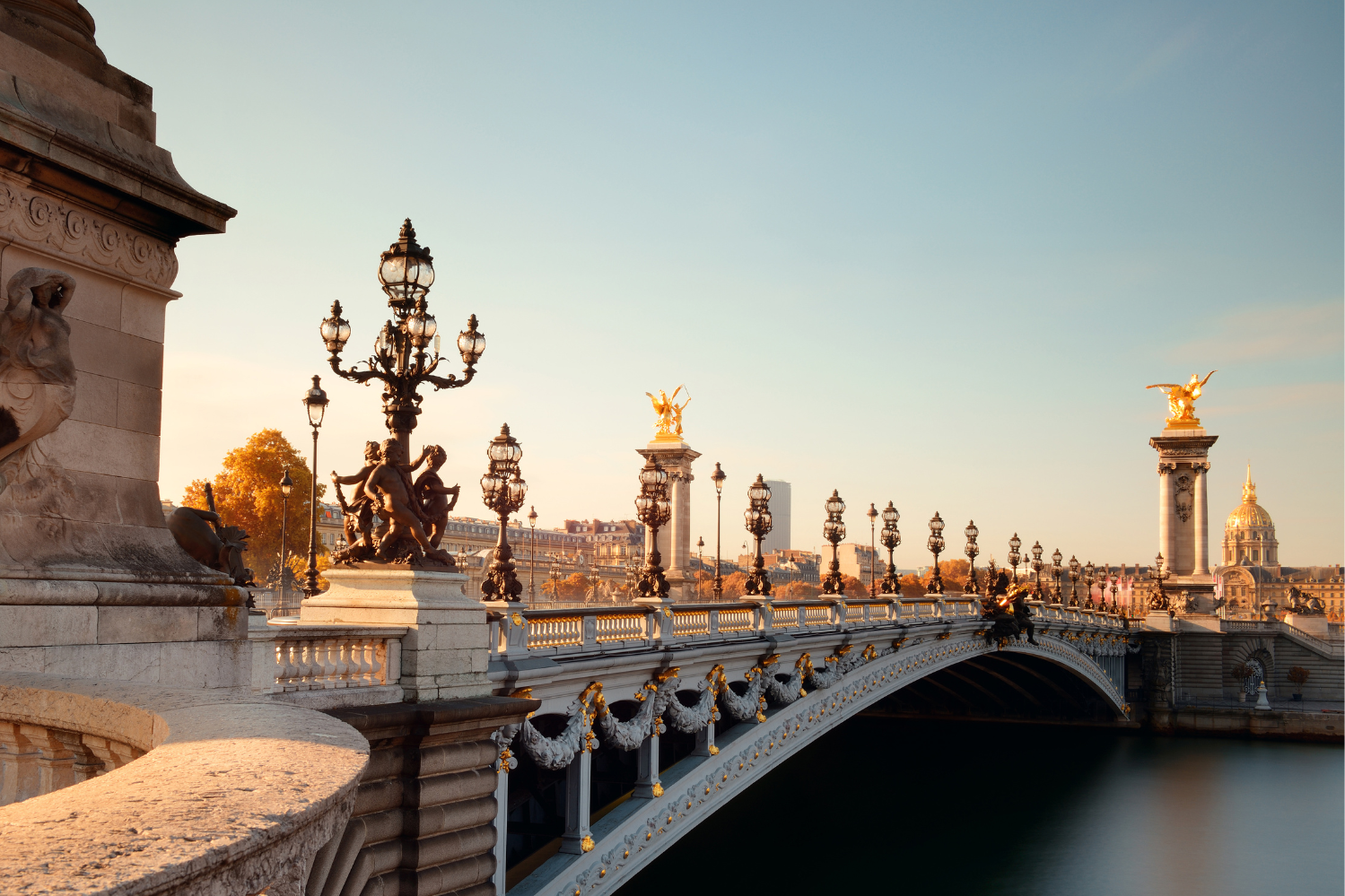 A stunning view of the Pont Alexandre III bridge in Paris during a golden hour. The ornate bridge features intricate sculptures, decorative lampposts, and gilded statues of winged horses on tall columns. In the background, the golden dome of Les Invalides shines under the warm sunlight, with autumnal trees adding a touch of color to the elegant cityscape.