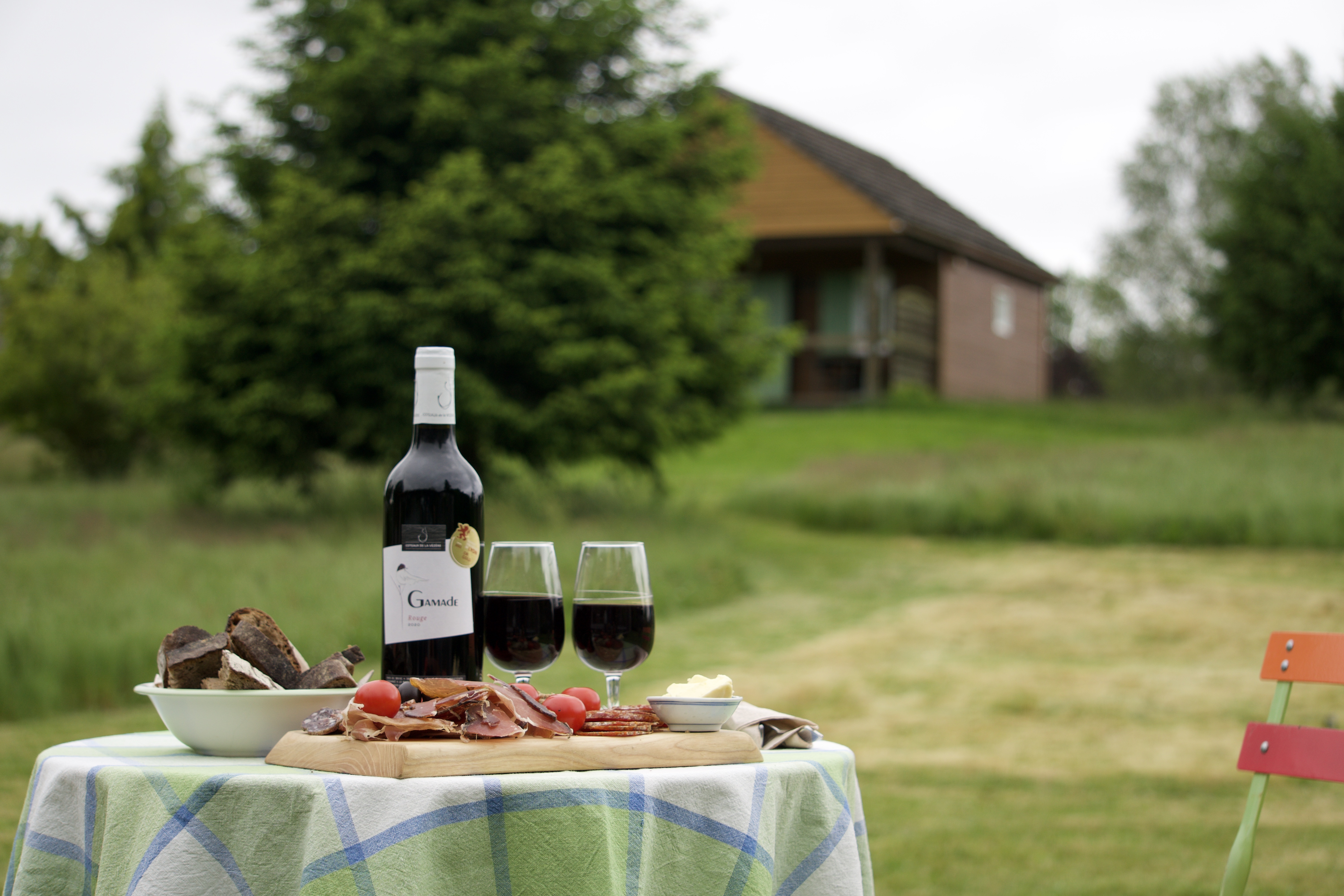 Bouteille et planche de charcuterie sur une table de jardin avec chalet et verdure en fond aux maisons de coline