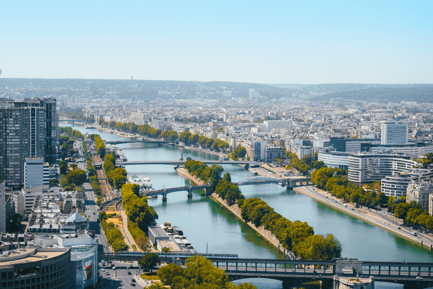 Une vue panoramique de Paris depuis les hauteurs, mettant en valeur la Seine avec ses nombreux ponts, des rues bordées d’arbres et des bâtiments modernes situés dans le 16ᵉ arrondissement, près du Parc André Citroën.