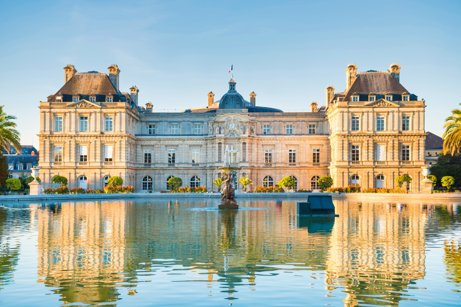 Le Palais du Luxembourg à Paris, reflété dans les eaux calmes de la fontaine du jardin, entouré de palmiers et de fleurs éclatantes par une journée ensoleillée.