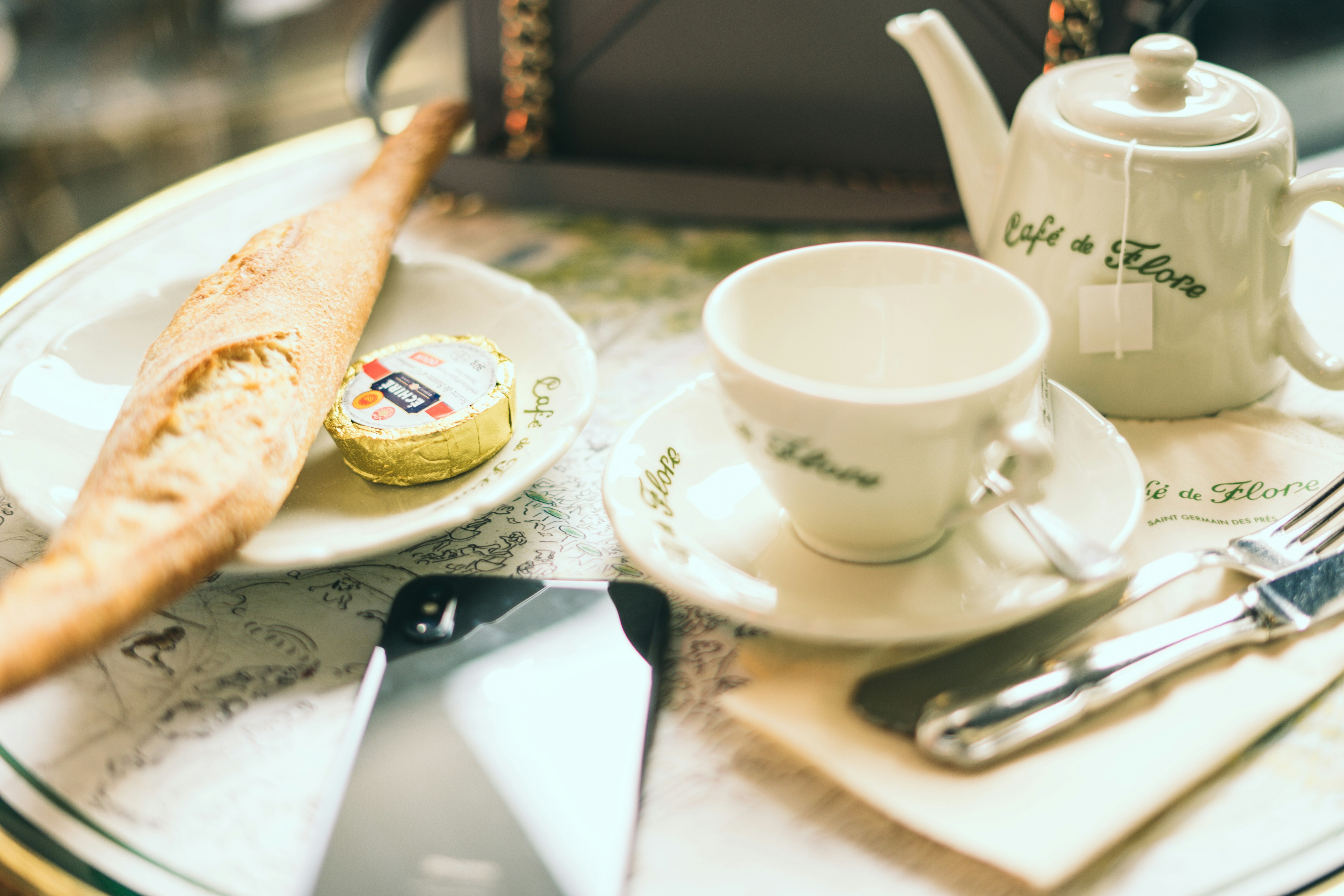 Petit-déjeuner au célèbre Café de Flore à Paris, avec une baguette, du beurre et du thé servis dans une vaisselle élégante aux couleurs de la maison, sur une table typique de café parisien.