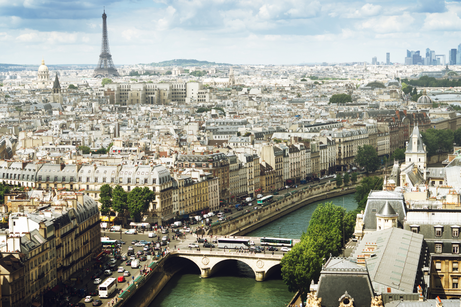 Vue panoramique de la Rive Gauche de Paris, avec la Tour Eiffel au loin, la Seine traversée par un pont historique, des bâtiments haussmanniens classiques et, à l’horizon, la silhouette moderne de La Défense.