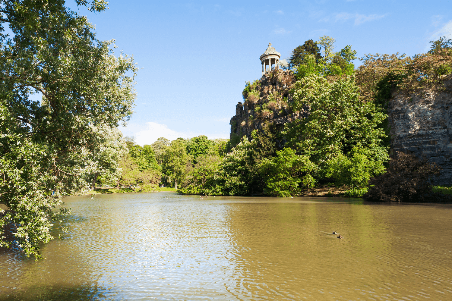 Parc des Buttes-Chaumont in the 19th arrondissement of Paris, featuring a serene lake, lush greenery, and the iconic Temple de la Sibylle atop its rocky hill, a perfect escape from the city's bustle.