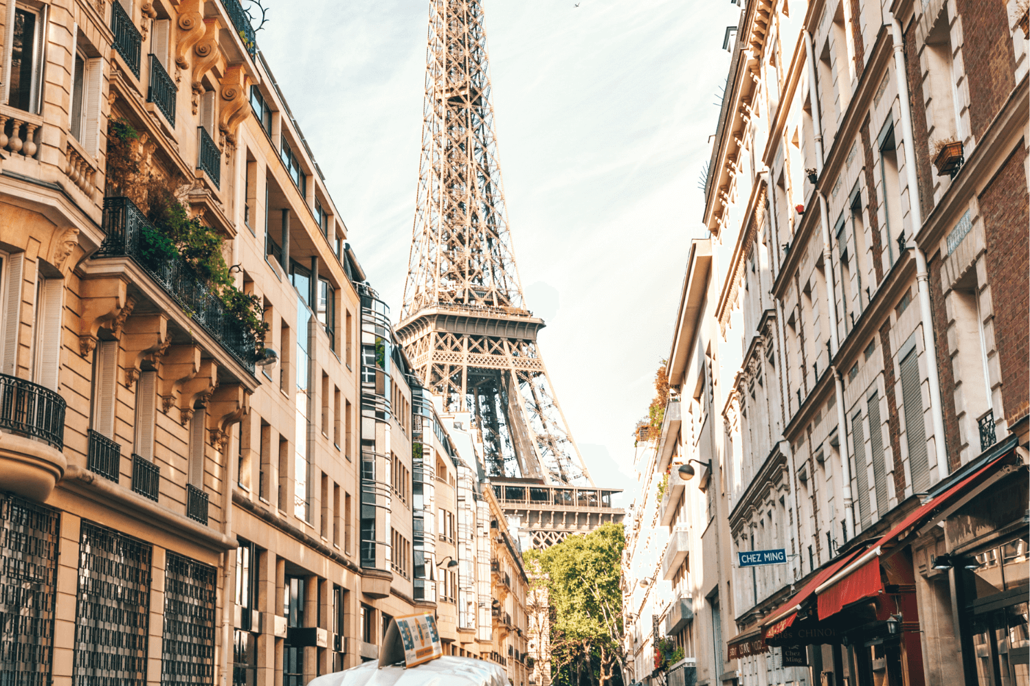 Street view in Paris with the Eiffel Tower in the background, surrounded by classic Haussmann-style buildings and modern architecture. A vibrant Parisian scene with shops, balconies, and warm sunlight enhancing the city's charm