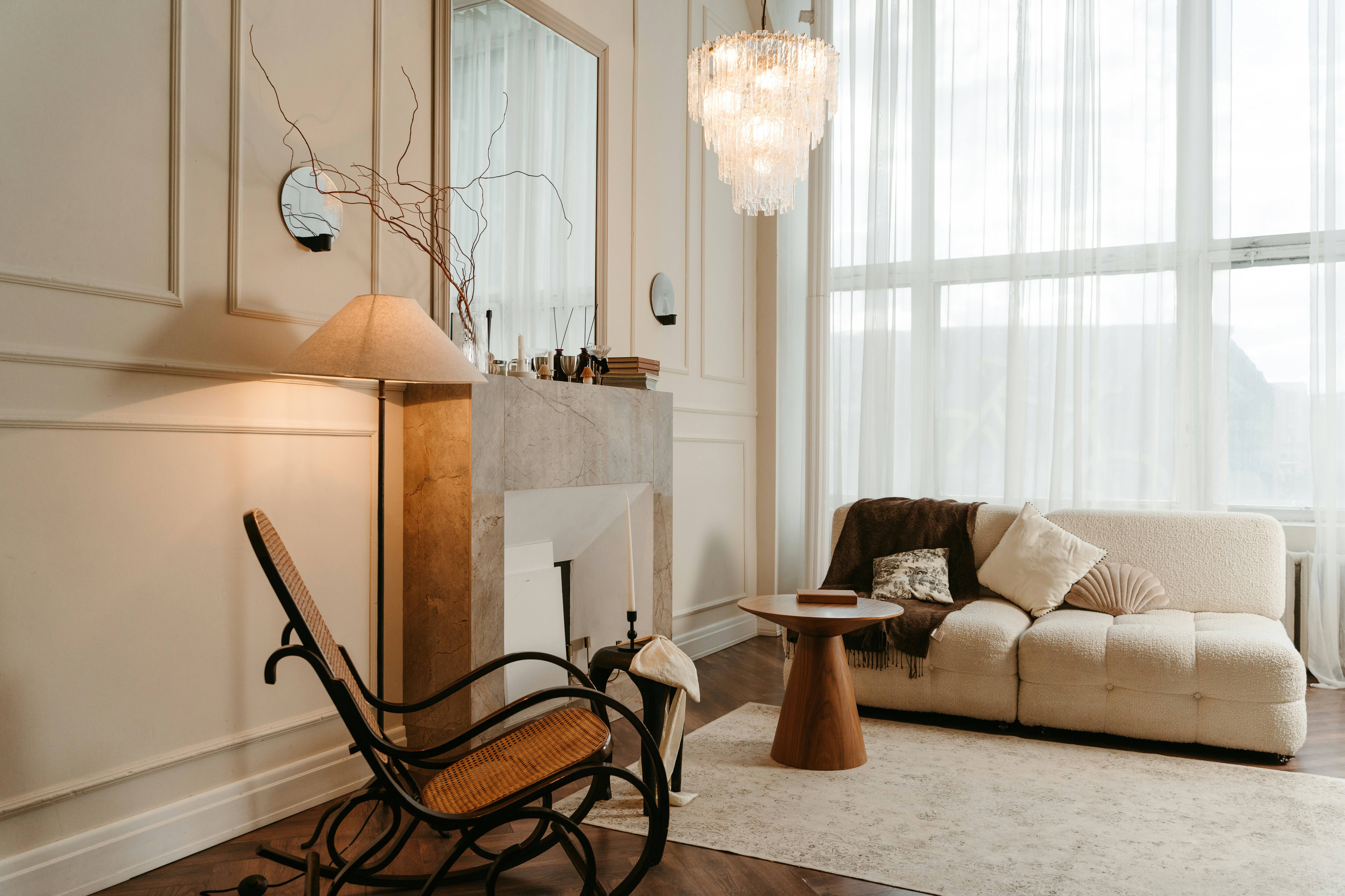 Cozy, elegant living room with a sofa, rocking chair, fireplace, and soft lighting by a large window.