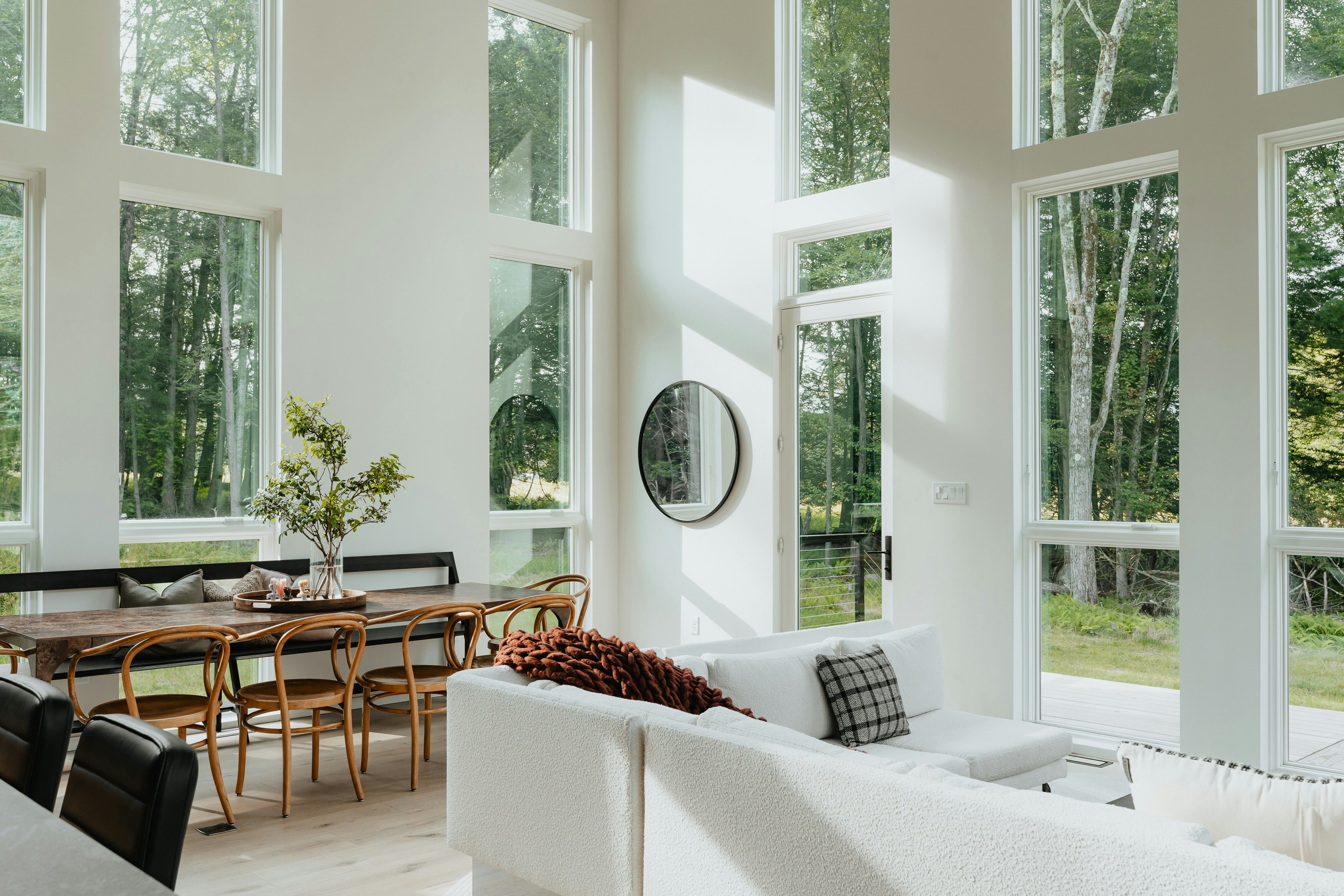 Bright modern living room with large windows, a light sofa, and a wooden dining table overlooking trees.
